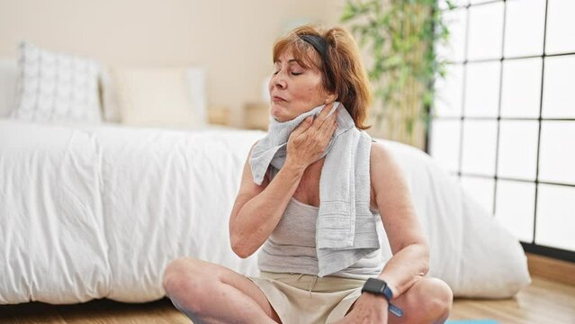 Middle age woman sitting on yoga mat sweating at bedroom