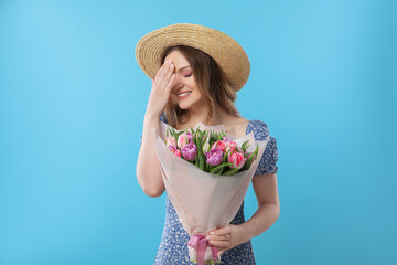 Happy young woman in straw hat holding bouquet of beautiful tulips on light blue background