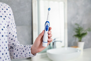 Woman holding electric toothbrush in bathroom at home, closeup