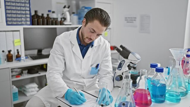 Young hispanic man scientist writing report smiling at laboratory