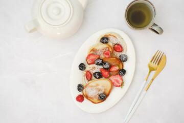 Pancakes with berries, green tea and teapot for breakfast on a light background.