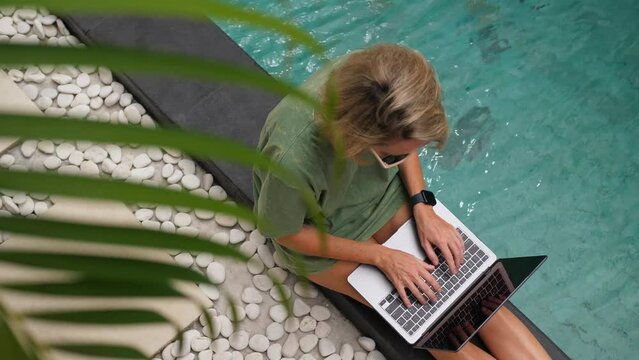 Female freelancer sitting near the swimming pool in tropical location, using laptop, top view. High quality 4k footage