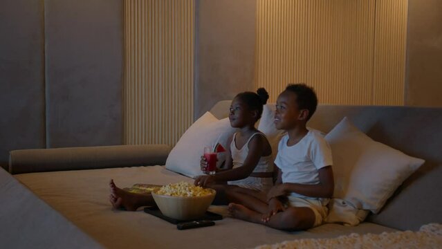 Two black children having good time resting in front of tv watching movie on cinema system, eating popcorn, family time concept, high quality