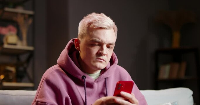 Frustrated young man uses smartphone. Angry dissatisfied annoyed irritated young hipster man aggressively scrolls through feed on phone, watches bad news, gets up throws smartphone on couch, leaves.