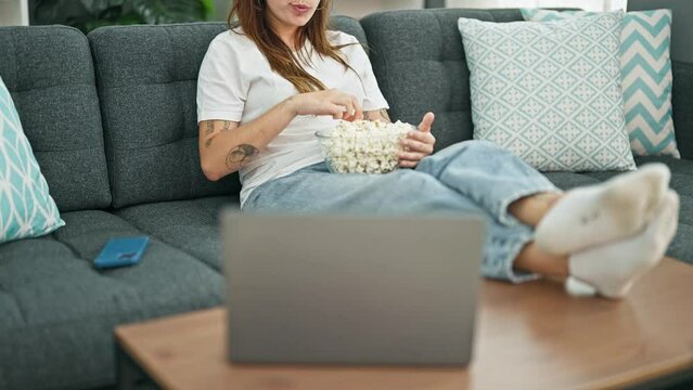 Young beautiful hispanic woman watching movie eating popcorn sitting on sofa at home