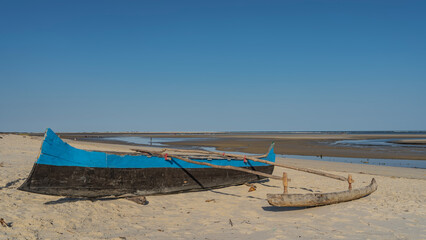 Homemade blue and black wooden boat on the sand at low tide of the ocean. Puddles of water are visible in the distance. Clear blue sky. Madagascar. Mozambique Channel. Morondava