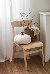 Seasonal autumn decor in scandinavian style interior . Pumpkin pillow, basket with a blanket and branches on a wooden chair in the living room