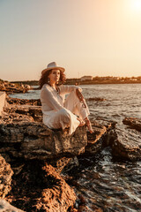 woman sea sunset. woman in a white pantsuit and hat is sitiing on the beach enjoying the sea. Happy summer holidays