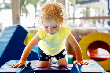 Happy blond little toddler girl having fun and sliding on indoor playground at daycare or nursery. Positive funny baby child smiling. Healthy girl climbing on slide.