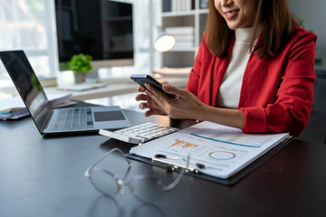 Asian businesswoman holding a smartphone using a calculator to calculate on graph paper Work record Budget calculations, taxes, financial accounts placed on the table in the office Management concept