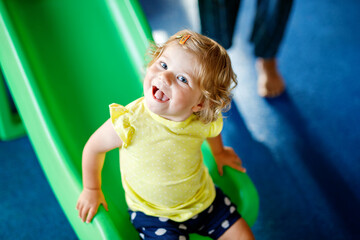Happy blond little toddler girl having fun and sliding on indoor playground at daycare or nursery....