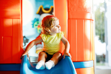 Happy blond little toddler girl having fun and sliding on indoor playground at daycare or nursery. Positive funny baby child smiling. Healthy girl climbing on slide. - obrazy, fototapety, plakaty