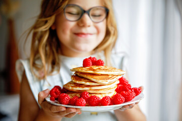 Little happy preschool girl with a large stack of pancakes and raspberries for breakfast. Positive child eating healthy homemade food in the morning.