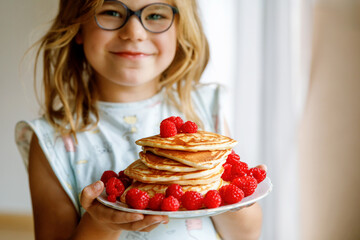 Little happy preschool girl with a large stack of pancakes and raspberries for breakfast. Positive...