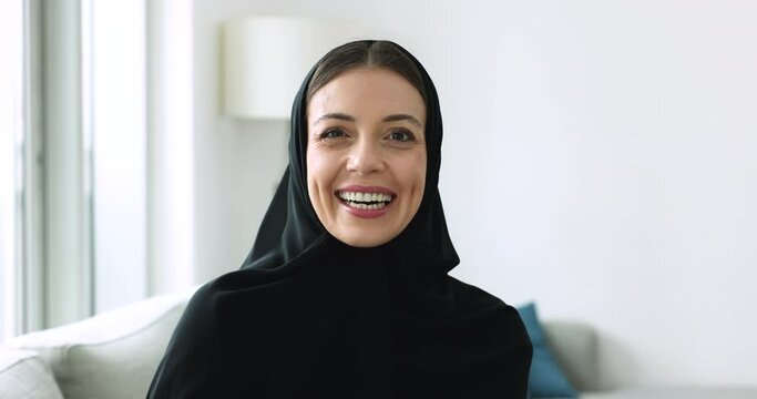 Attractive Muslim woman in black hijab sit on sofa indoors. Portrait of happy middle-aged 45s female wear traditional Islamic robe with scarf smile looks at camera resting at home. Culture, lifestyle