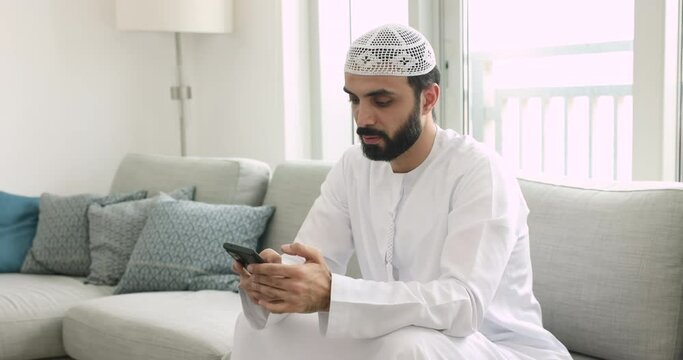 Serious Muslim man in traditional clothes using smartphone at home, share messages, solve business distantly, typing e-mail, contact to client remotely by correspondence, using new mobile app. Tech