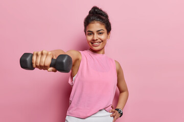 Photo of young motivated Iranian girl with dark hair holds dumbbell and smiles pleasantly goes in for sport regularly dressed in activewear isolated over pink background leads fit active lifestyle