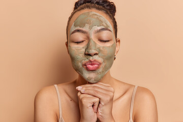 Photo of pretty young woman with dark hair keeps hands under chin lips folded eyes closed applies beauty clay mask on face wears t shirt isolated over brown background. Facial treatments concept