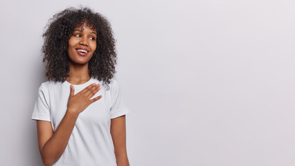 Horizontal shot of beautiful curly haired woman keeps hand on chest feels thankful and delighted smiles pleasantly dressed in casual t shirt isolated over white background empty space for your advert