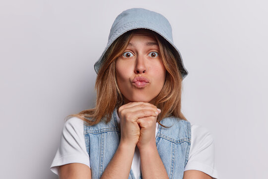 Studio shot of lovely adult European woman keeps lips rounded hands under chin concentrated at camera wears denim panama and vest isolated over white background. Human facial expressions concept
