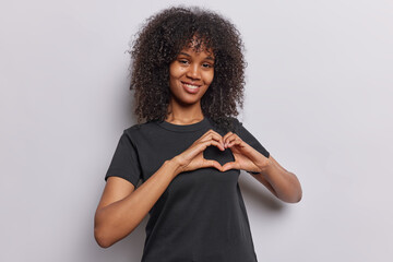 Horizontal shot of friendly black woman with curly hair makes love gesture expresses kindness dressed in casual black t shirt isolated over white background. Romantic female model shows heart sign - obrazy, fototapety, plakaty