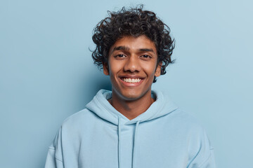 Horizontal shot of handsome curly haired Hindu man dressed in casual sweatshirt smiles gladfully focused at camera attentively isolated over blue background. People and positive emotions concept