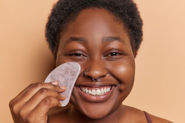 Close up shot of positive dark skinned woman uses gua sha stone for massaging face smiles broadly...