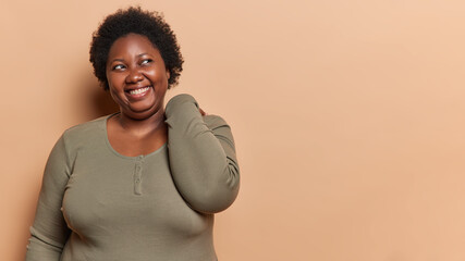 Lovely dark skinned plus size young woman keeps hand on neck smiles broadly shows white teeth...