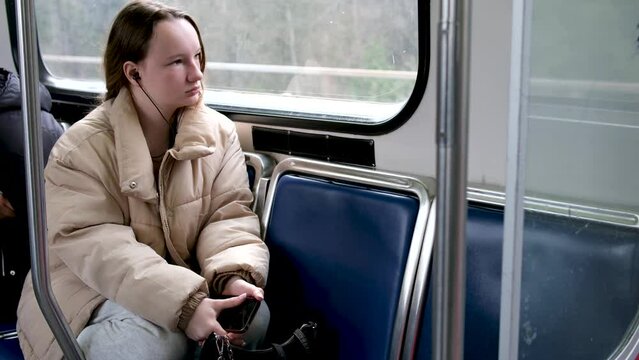 teenage girl rides in skytrain on train in bus headphones in ears listens to music autumn spring winter warm beige jacket hair is tied down real person sits tucked legs under sad dreams awaits stop