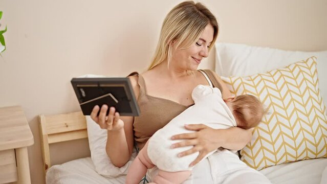Mother and daughter sitting on bed breastfeeding baby looking photo at bedroom