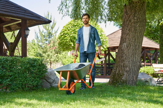 Attractive male landscaper pushing metal wheelbarrow, while working in garden. Low angle view of smiling gardener in jeans using cart for transportation, while walking on grass. Concept of gardening. - Powered by Adobe