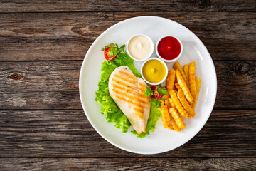 Grilled chicken breast, French fries and fresh vegetables on wooden table
