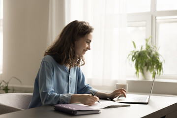Cheerful freelancer girl working from home, using laptop, writing notes, consulting online service,...