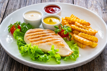 Grilled chicken breast, French fries and fresh vegetables on wooden table
