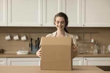 Happy beautiful young customer woman opening package, unpacking cardboard box, parcel on kitchen table, looking inside, smiling, getting delivered purchase from Internet shop, grocery store