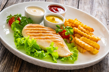 Grilled chicken breast, French fries and fresh vegetables on wooden table
