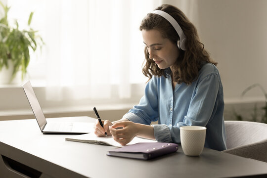 Modern gen Z young adult student girl in earphones studying on Internet at laptop, listening to educating audio lecture, webinar, writing notes, sitting at home office work table