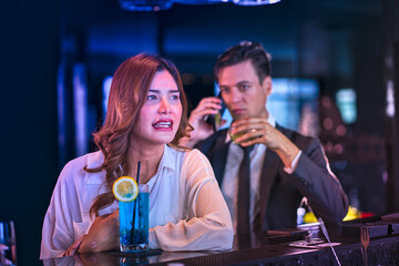 Young pretty asian girl at bar drinking cocktail and a businessman behind with cell phone.