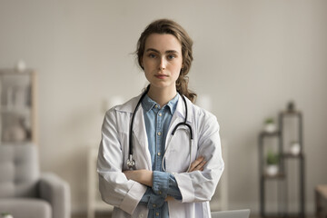 Serious confident pretty young doctor woman in white uniform coat posing with arms folded, looking...