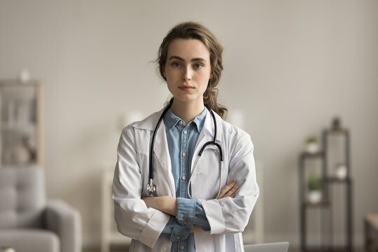 Serious confident pretty young doctor woman in white uniform coat posing with arms folded, looking at camera. Female practitioner, medical worker professional head shot portrait