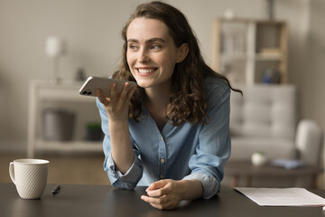 Cheerful gen Z young entrepreneur girl discussing business project on mobile phone at home office table, talking on speaker, speaking at gadget, recording voice audio message