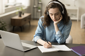 Serious pensive online student girl in headphones listening to audio lesson, learning course on laptop, writing notes in notebook, using Internet technology for education, studying at home