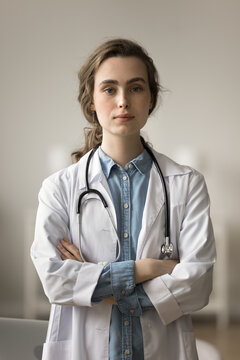 Serious medical intern, beautiful young doctor woman vertical portrait. Female practitioner with stethoscope on neck looking at camera, posing, standing with hands crossed