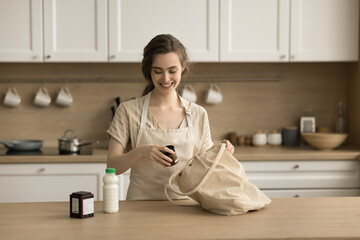 Positive pretty young woman in apron unpacking shopping bag in kitchen, getting grocery purchase,...