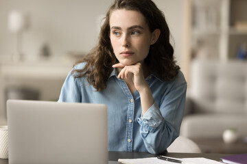 Obraz premium Serious thoughtful young gen Z freelance employee girl thinking on online business project, sitting at laptop at home workplace, looking away, leaning chin on hand in deep thoughts