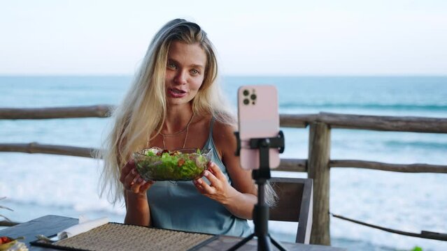 Food blogger woman films video about green salad bowl, dieting for fit skinny body at cafe, sea view. Travel influencer tells about local cuisine, diet on social network. Vegan female shoots vlog.