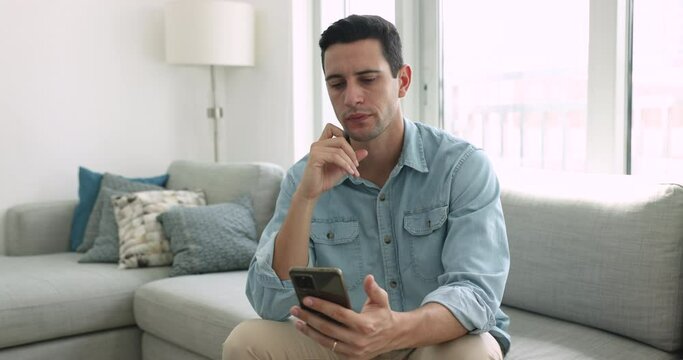 Young serious Hispanic man holding smartphone seated on couch, looks at modern gadget screen, read notification or message feels thoughtful, ponder over answer, consider received information by e-mail