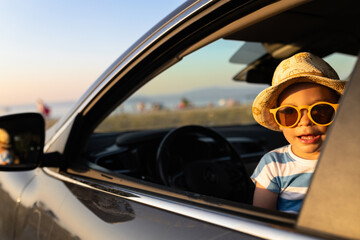 Fashionable baby boy, peeks out of the car in the sunset. Beach and sea in the background. Travel summer vacation concept.