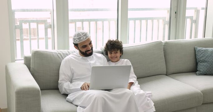 Happy Muslim man with son watching amusing on-line content on laptop, smile look at device screen discuss new video in social media, make order use electronic commerce web site, spend time on internet