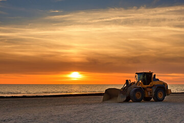 Fototapeta premium Bulldozer on the beach at sunset. A large tractor cleans the beach of sea grass. Bulldozer in motion and blur.